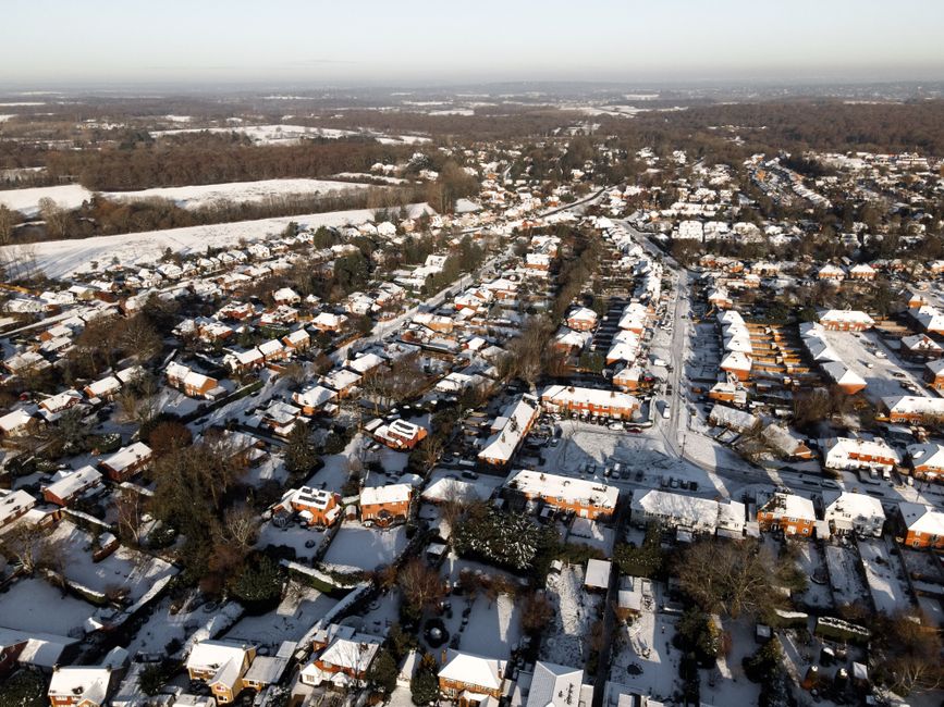 An aerial view of a residential neighbourhood in winter, showing numerous single-family houses with snow-covered roofs, arranged along curving streets and lanes. The houses have brick walls and tiled